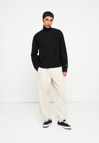 Suéter negro de punto con cuello alto, con puños y dobladillo acanalados, combinado con pantalones crema de corte holgado y zapatillas negras con cordones blancos.