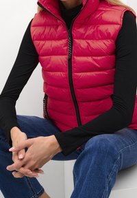 Czerwona pikowana kamizelka z wysokim kołnierzem i zamkiem błyskawicznym, noszona na czarnym topie z długim rękawem. Modelka siedzi z splecionymi rękami, widoczne dżinsy.