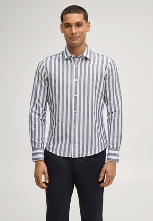 Mann steht und trägt ein langärmliges Hemd mit vertikalen grauen und weißen Streifen sowie eine dunkelblaue Hose.
