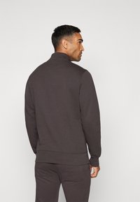 Mørkegrå hettegenser med høy krage og ribbestrikkede mansjetter. Mykt stoff med et strømlinjeformet design, som har minimale sømdetaljer.