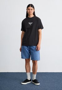 Sort bomulds t-shirt med hvid "Porsche League Turbo Club" grafik, parret med lyseblå denimshorts, sorte sneakers og hvide sokker.