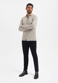 Lichtgrijs lang shirt met knopen aan de voorkant, gecombineerd met zwarte slim-fit broek en zwarte enkellaarsjes. Soepele textuur, op maat gemaakt.