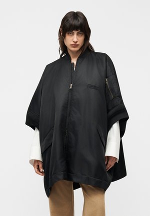 Femeie cu păr închis la culoare purtând o pelerină neagră supradimensionată cu fermoar peste mâneci albe și pantaloni bej, stând în fața unui fundal simplu, deschis la culoare.