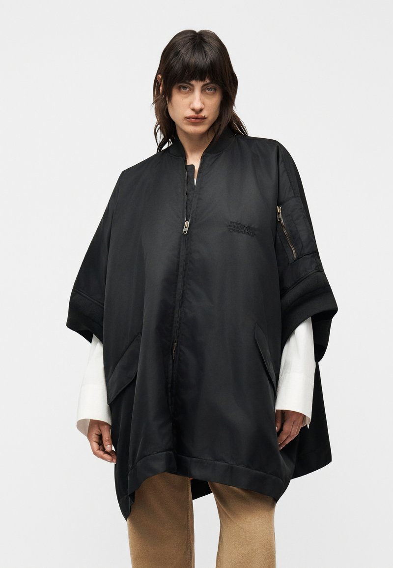 Femme aux cheveux bruns portant une cape noire zippée oversize sur des manches blanches et un pantalon beige, debout devant un fond clair uni.