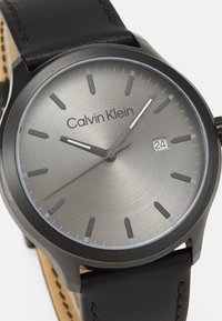 Reloj Calvin Klein con un dial metálico gris, marcadores de hora negros y una pantalla de fecha. Con una correa de cuero negro con costuras visibles.