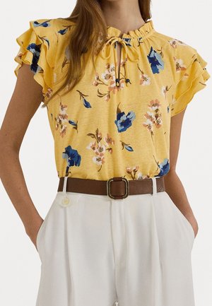 Gele bloemenblouse met gerimpelde korte mouwen, gebonden keyhole-halslijn, ingestopt in hooggetailleerde witte broek met een bruine riem met gouden gesp.