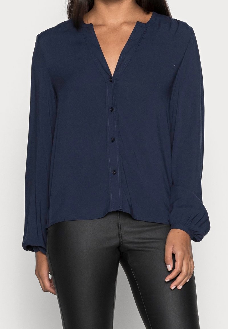 Femme portant un chemisier bleu marine à manches longues avec boutons et un pantalon en cuir noir, debout sur un fond clair.