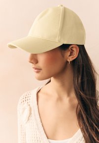 Light yellow baseball cap made of textured fabric, featuring a curved brim and stitched detailing, worn casually at an angle.