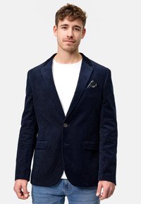 Marineblå kordfløyelsblazer med notched lapels, enkel knappelukking, to frontlommer og et mønstret lommetørkle. Båret over en hvit skjorte.