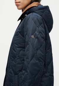 Μπλε ναυτικό καπιτονέ μπουφάν με κουκούλα, με καφέ γιακά και κεντημένο λογότυπο "RJL" στο αριστερό μανίκι. Λεία υφή, μήκος μέχρι τη μέση.