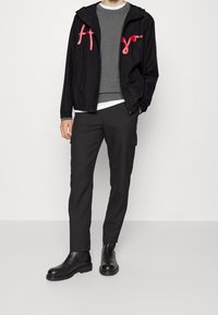 Black windbreaker jacket with pink logo accents, grey sweater underneath, black cargo pants, and black ankle boots. Smooth fabric, relaxed fit.