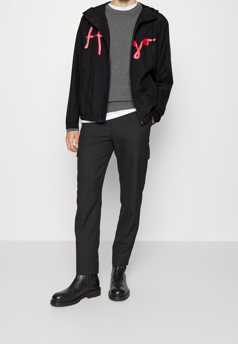 Black windbreaker jacket with pink logo accents, grey sweater underneath, black cargo pants, and black ankle boots. Smooth fabric, relaxed fit.