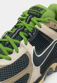 Παπούτσι Nike Vomero 5 με επάνω μέρος από πλέγμα σε γκρι με μπεζ λεπτομέρειες, φωτεινές πράσινες κορδόνες και μαύρο λογότυπο swoosh. Λείο, υφασμάτινο πέλμα.
