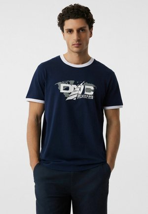 Mężczyzna w granatowej koszulce z białym obszyciem i logo żaglówki z napisem "Diverse Nautical Club", ręce w kieszeniach, neutralne tło.