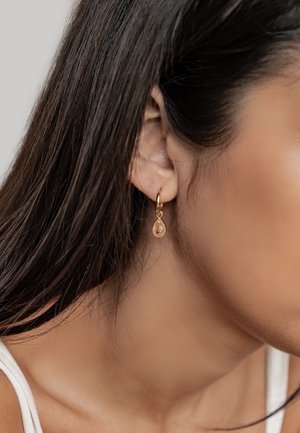 Goldfarbene Creolen mit einem tropfenförmigen Bernstein-Anhänger, der mit einer strukturierten Akzentuierung um den Stein versehen ist.