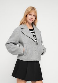 Νεαρή γυναίκα με γκρι μάλλινο σακάκι με μεγάλους γιακάδες, ριγέ πουκάμισο και μαύρη φούστα, στέκεται με τα χέρια στις τσέπες μπροστά σε λευκό φόντο.