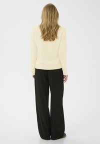 Kvinna med l�ångt blont hår, klädd i en krämfärgad tröja och svarta vida byxor, stående med ryggen mot kameran på en vit bakgrund.