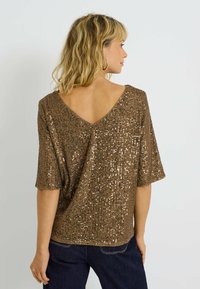 Top de lentejuelas bronce con escote en V y mangas cortas, que presenta una superficie texturizada y un corte holgado, combinado con unos jeans de mezclilla oscura.