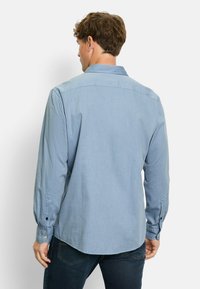 Mann, der von hinten schaut, bekleidet mit einem hellblauen Langarmshirt und dunklen Jeans vor einem schlichten weißen Hintergrund.