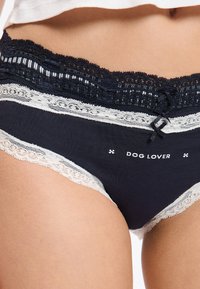 Mutandine di cotone blu scuro con bordo in pizzo bianco e stampa "DOG LOVER". Presentano un fiocco decorativo sulla fascia in vita.