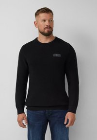 Homme aux cheveux courts et à la barbe, portant un pull en maille noir et un jean bleu, se tenant devant un fond gris clair uni.