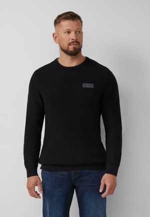 Uomo con capelli corti e barba, che indossa un maglione nero a trama e jeans blu, in piedi davanti a uno sfondo grigio chiaro semplice.