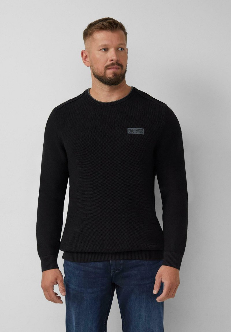 Homme aux cheveux courts et à la barbe, portant un pull en maille noir et un jean bleu, se tenant devant un fond gris clair uni.