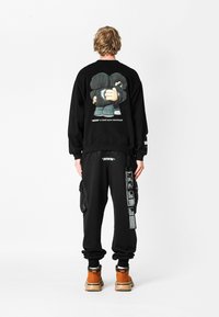 Sudadera negra con un diseño gráfico en la parte posterior, combinada con pantalones joggers negros que tienen bolsillos tipo cargo y detalles en texto. Calzado marrón.