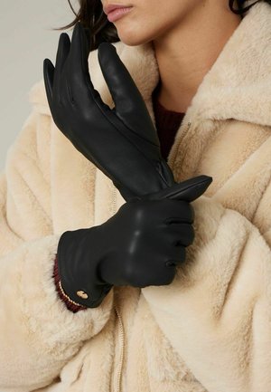 Personne portant des gants en cuir noir et un manteau en fausse fourrure beige, ajustant un gant sur l'autre main devant un fond neutre.