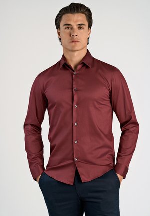 Man standing with hands in pockets wearing a buttoned maroon long-sleeve shirt and dark pants against a plain background.
