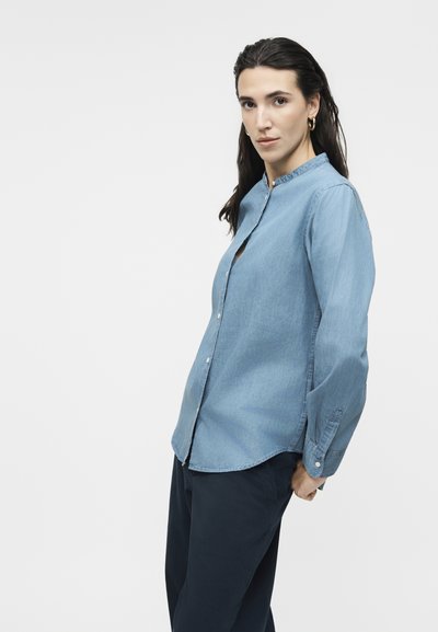 Γυναίκα με μακριά σκούρα μαλλιά, που φοράει χρυσούς κρίκους σκουλαρίκια, γαλάζιο πουκάμισο με κουμπιά και σκούρο παντελόνι, μπροστά σε απλό φωτεινό φόντο.