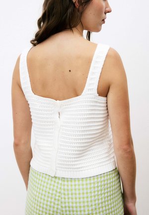 Crop top en tricot blanc avec des bretelles larges, présentant un côtes texturé et une fermeture à boutons au dos. Associé à un pantalon à carreaux verts.