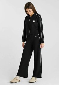 Zwarte cropped hoodie met witte zijkanten en logo, gecombineerd met zwarte wijde broek met een bijpassend logo en zijkanten.