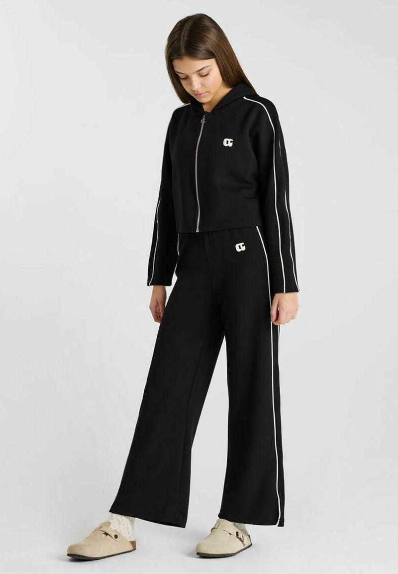 Zwarte cropped hoodie met witte zijkanten en logo, gecombineerd met zwarte wijde broek met een bijpassend logo en zijkanten.