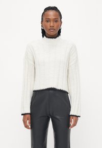 White ribbed knit sweater with a high neck and cropped hem, featuring black trim along the edges. Paired with black faux leather pants.