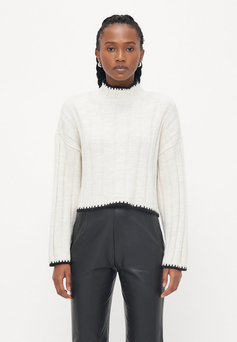 White ribbed knit sweater with a high neck and cropped hem, featuring black trim along the edges. Paired with black faux leather pants.