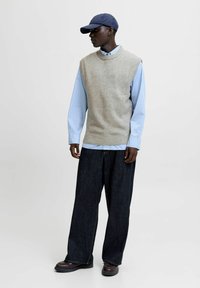 Un gilet en maille gris clair sur une chemise à rayures bleues, associé à un pantalon en denim large de couleur foncée et une casquette marine, posé sur un fond neutre.