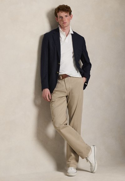 Fiatal férfi, aki a falnak dől, tengerészkék blézert, fehér galléros pólót, bézs nadrágot, barna övet és fehér tornacipőt visel, kezével a zsebében.