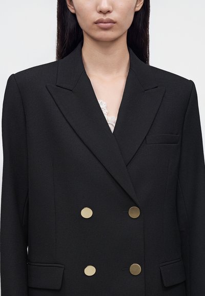 Femme portant un blazer noir croisé à quatre boutons dorés et un haut blanc orné de dentelle en dessous, montrée des épaules à la taille.