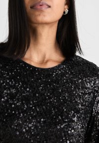 Top preto com lantejoulas, de decote arredondado, adornado com lantejoulas brilhantes densamente empilhadas que criam uma superfície texturizada. Acessórios são visíveis.