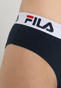 Čierne pánske nohavičky z bavlny s bielym pásikom, na ktorom je logo Fila v čiernej a červenej farbe. Hladký materiál, priliehavý strih, zakrivené otvory na nohách.