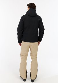 Juodas izoliuotas džemperis su gobtuvu; turi elastingas rankoves ir logotipo detalę. Sujungtas su šviesiai rudos spalvos sniego kelnėmis, kurios demonstruoja laisvą pristatymą ir batus.