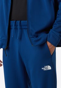 Blauwe sportjack en broek gemaakt van een gestructureerde stof, met een ritssluiting aan de voorkant en een logo op het broekbeen. Trekkoorden in de taille.