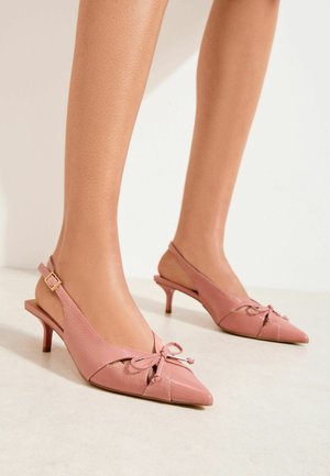 Nohy v ružových lodičkách so špičatou špičkou a opätkom na remienku s malými mašličkami, stojace na svetlej podlahe na jednoduchom pozadí.