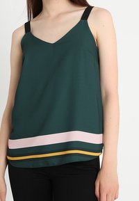 Zelený tílko bez rukávů s výstřihem do V, černými ramínky a horizontálními pruhy v růžové, tmavě zelené a žluté na okraji. Hladký povrch.