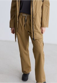 Braune wasserdichte Jacke mit passender Hose. Verstellbare Kordelzug an der Hose, Reißverschluss vorne. Glatte Textur, lockere Passform und knöchellang.