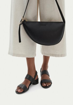 Borsa a tracolla a mezzaluna in pelle nera indossata con pantaloni beige a gamba larga e sandali neri con tacco largo su una persona dalla pelle scura.