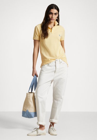 Yellow polo shirt, white relaxed jeans, and white sneakers. Beige tote bag with blue accents. Person stands with hands on hips.