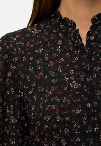 Blouse noire en tissu texturé avec un motif floral dans les tons bordeaux, blanc et vert. Col volanté et détail de fermeture à boutons.