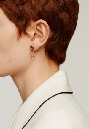 Pendiente de tono dorado con una gran piedra de ámbar, con cadenas colgantes y pequeñas cuentas. Textura suave, diseño moderno y caída elegante.